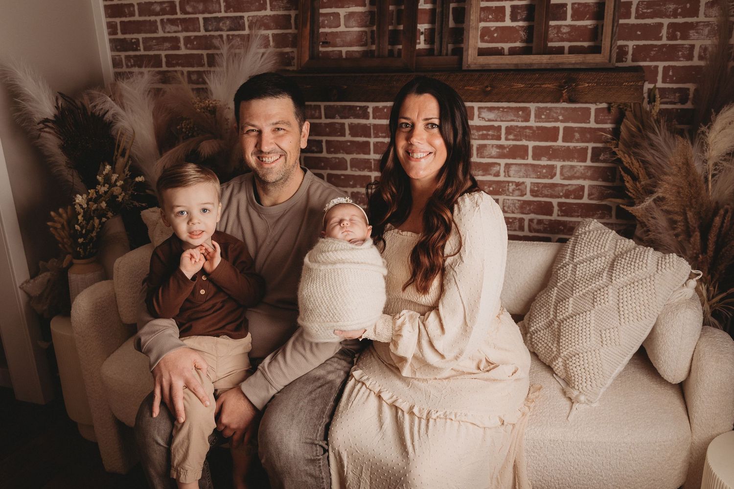 A happy family of four with a toddler and swaddled newborn poses on a couch in a warm studio.