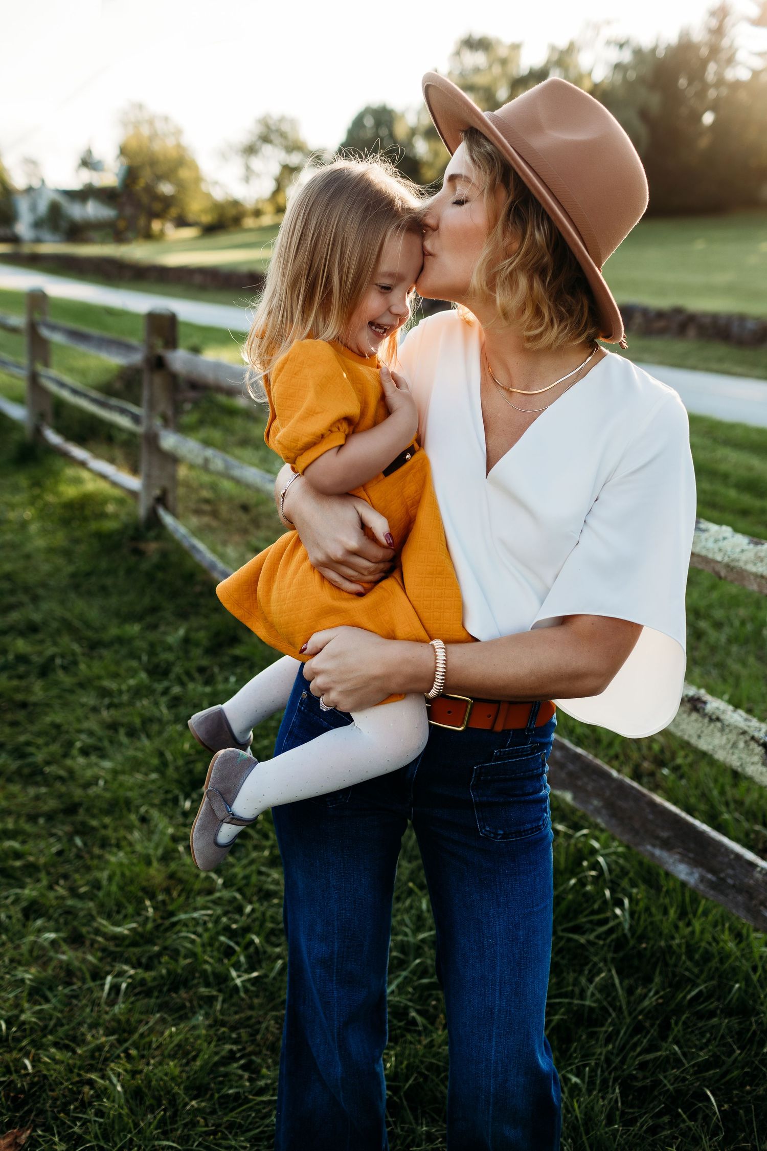 Mother in hat and white top kissing young daughter in orange dress outdoors near fence in Newtown Square, pa