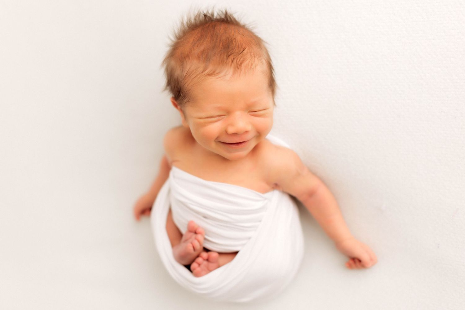 A newborn resting peacefully in a white wrap on a soft white background with a sweet smile.