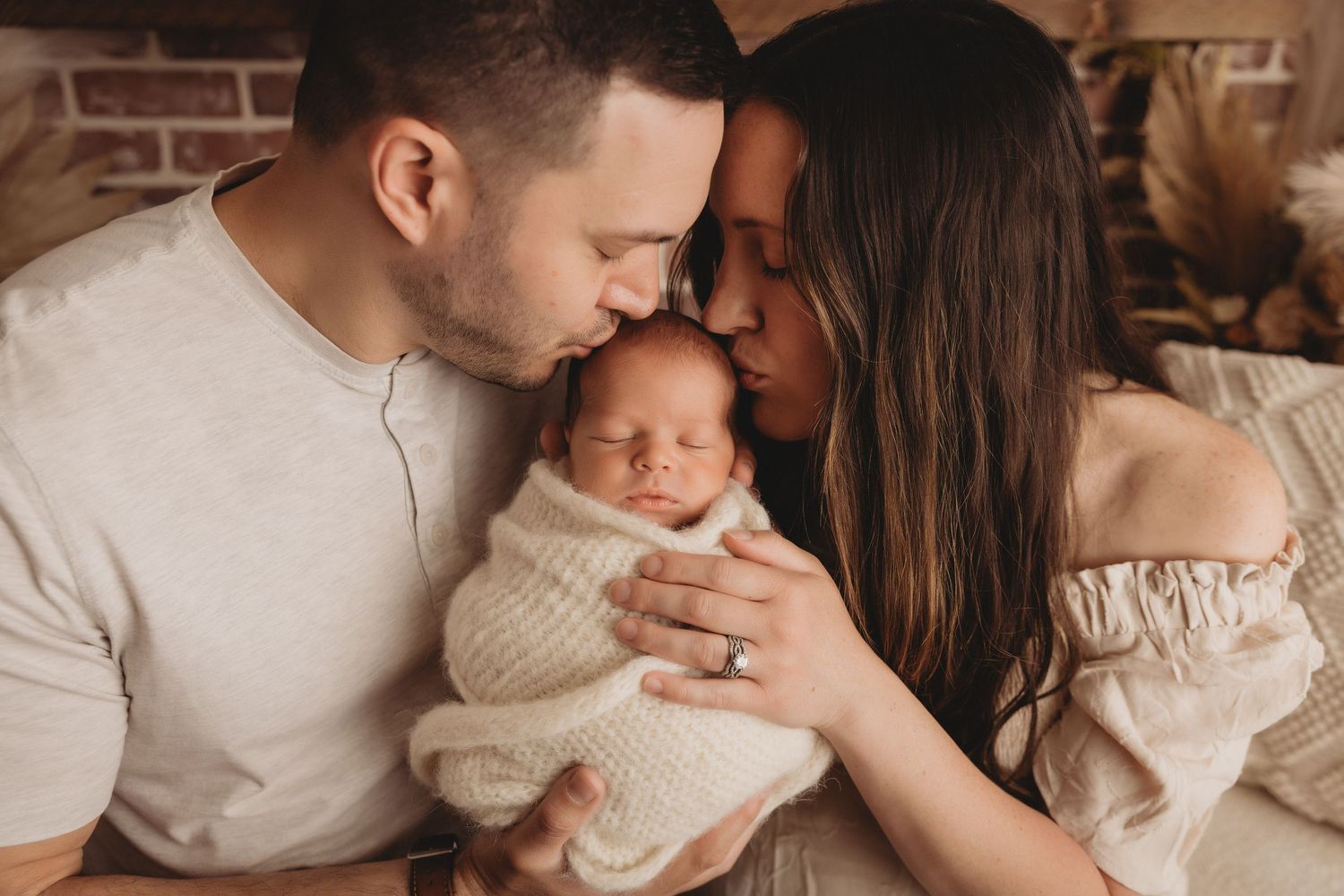 New parents tenderly touch foreheads over their sleeping newborn baby wrapped in white.