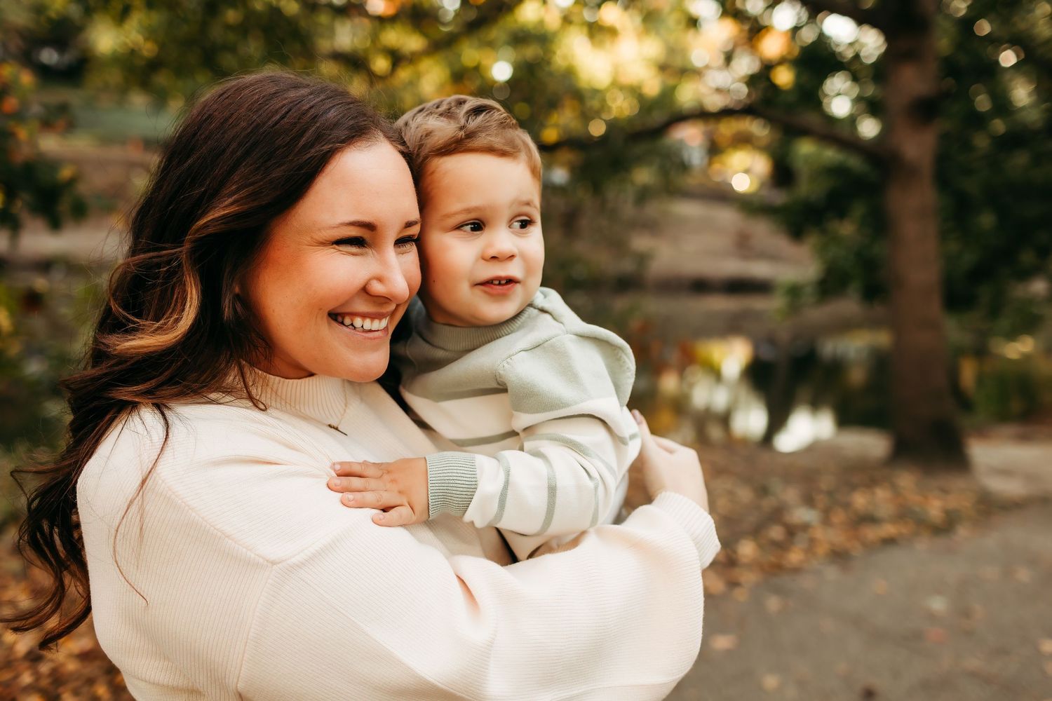 Smiling mother holding young toddler boy in green hoodie in autumn outdoor portrait.