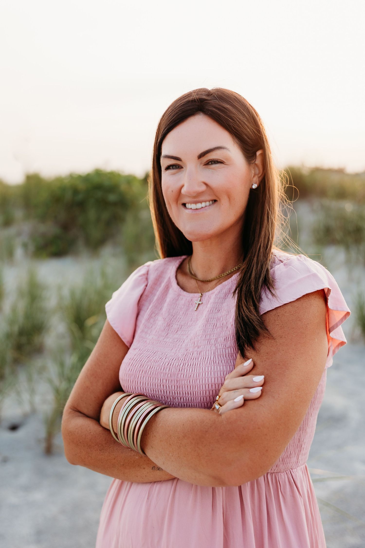 Smiling woman in pink dress with arms crossed poses on sandy beach with dunes and greenery in background.