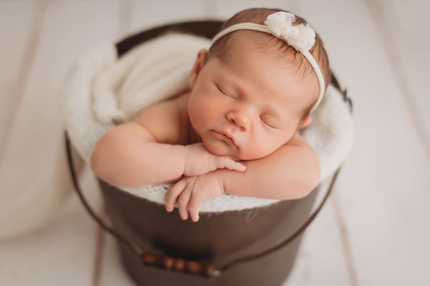 Sleeping newborn baby in a brown bucket with white fleece lining, wearing a delicate white bow headband.