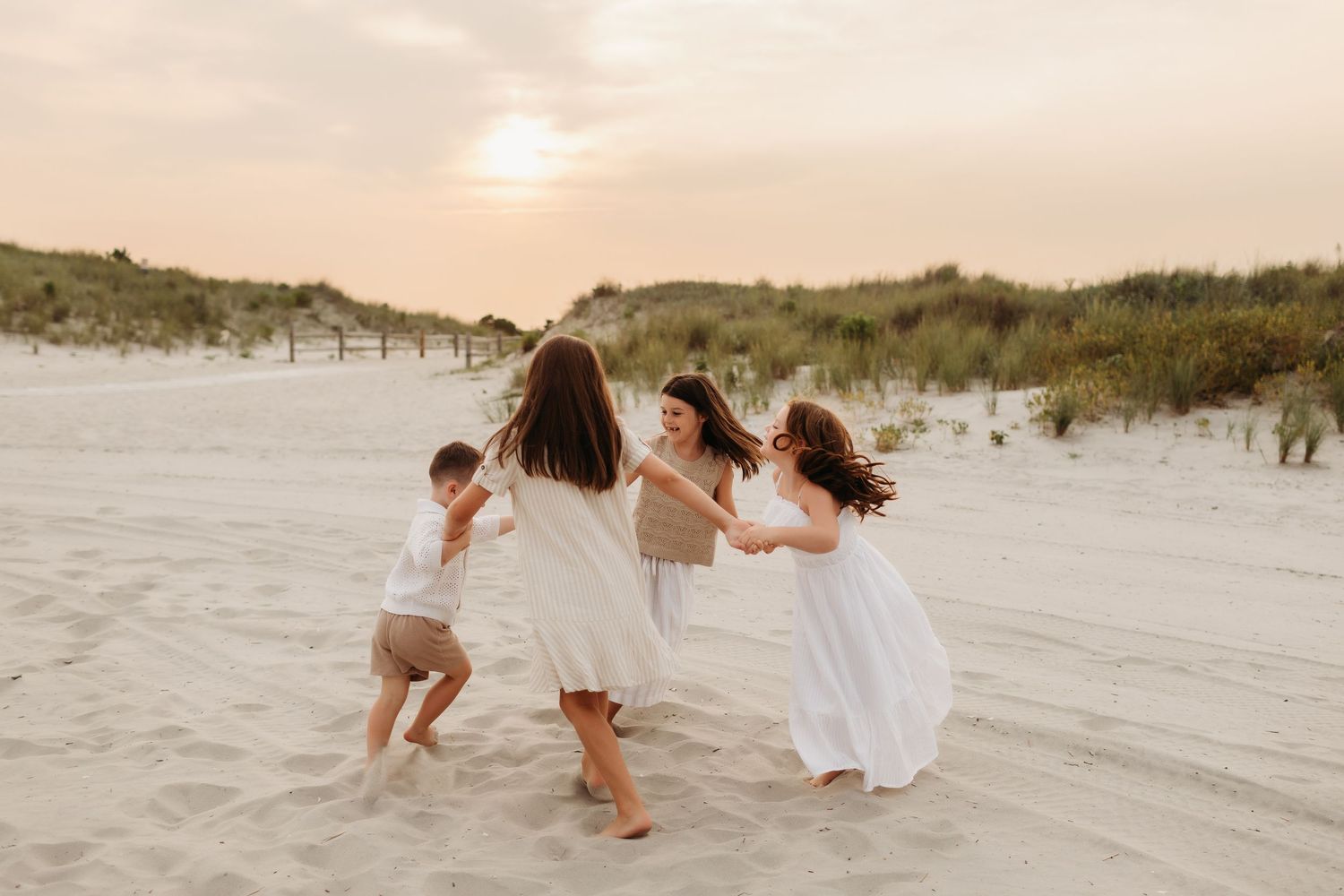 A joyful family plays together on a sandy beach at sunset, with children spinning around their mother in white dresses.