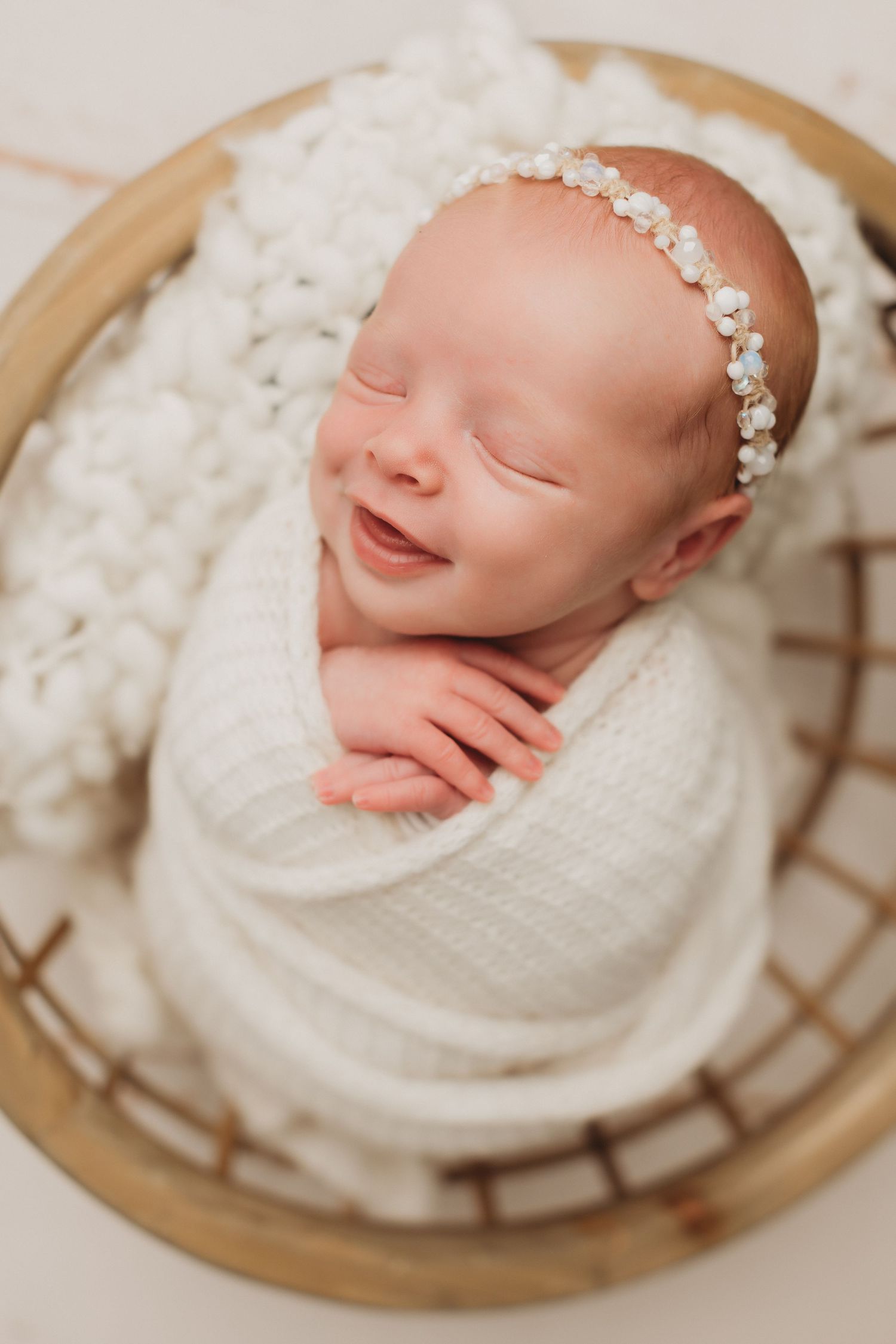 Smiling newborn baby swaddled in white wrap wearing floral headband sleeping in a wicker basket.