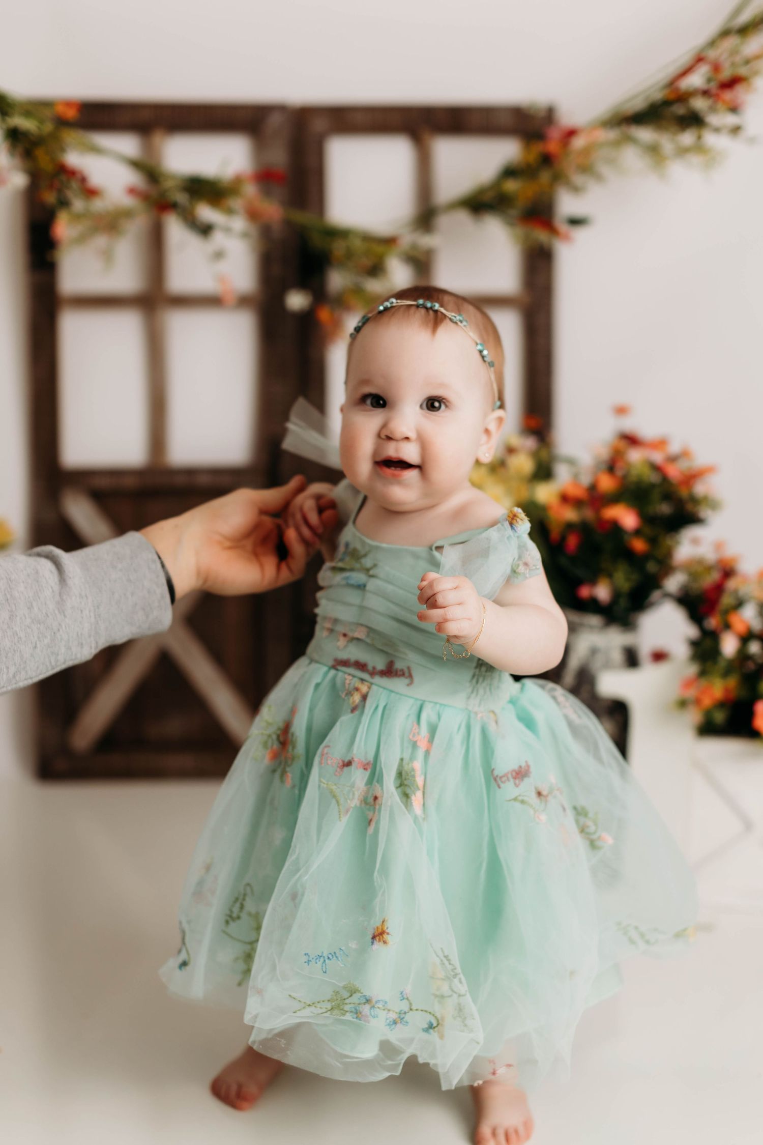 A small child wears a mint green tulle dress with floral details in front of a rustic window backdrop with orange flowers.