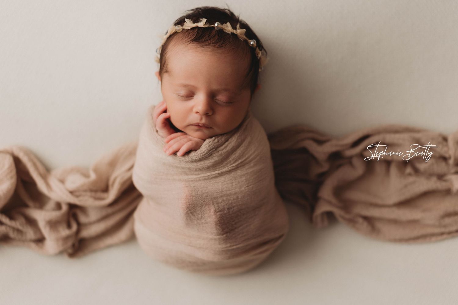 A sleeping newborn baby swaddled in beige cloth with a floral headband on a white background.