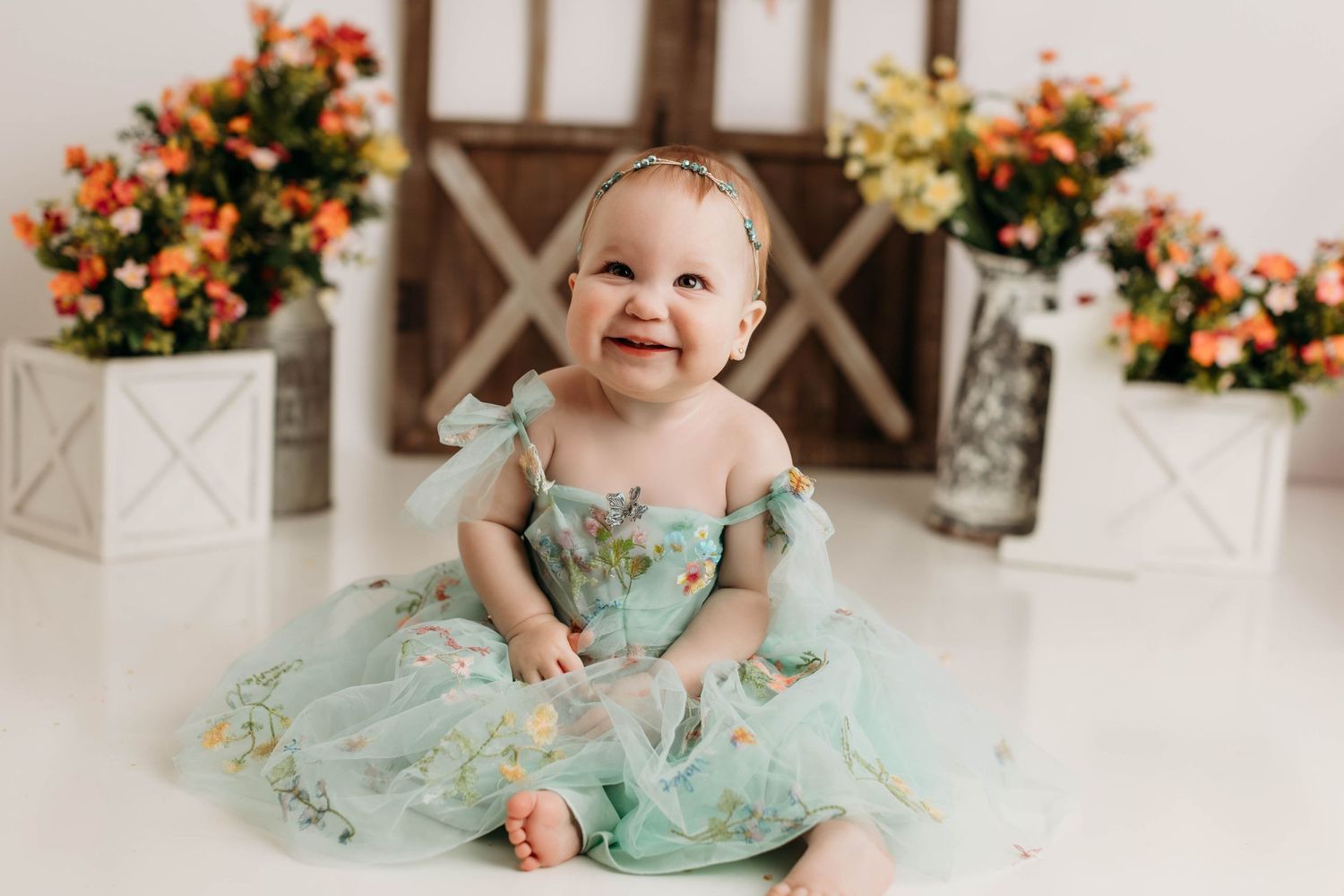 A small child sits on a white floor wearing a mint green tulle dress, surrounded by orange and yellow floral arrangements.