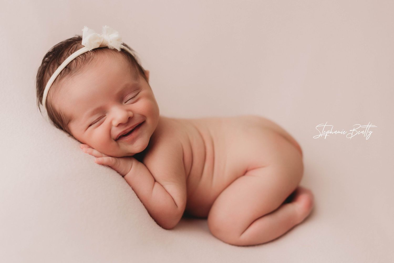 Newborn baby wearing white headband smiles while lying on soft pink backdrop during professional photo session.