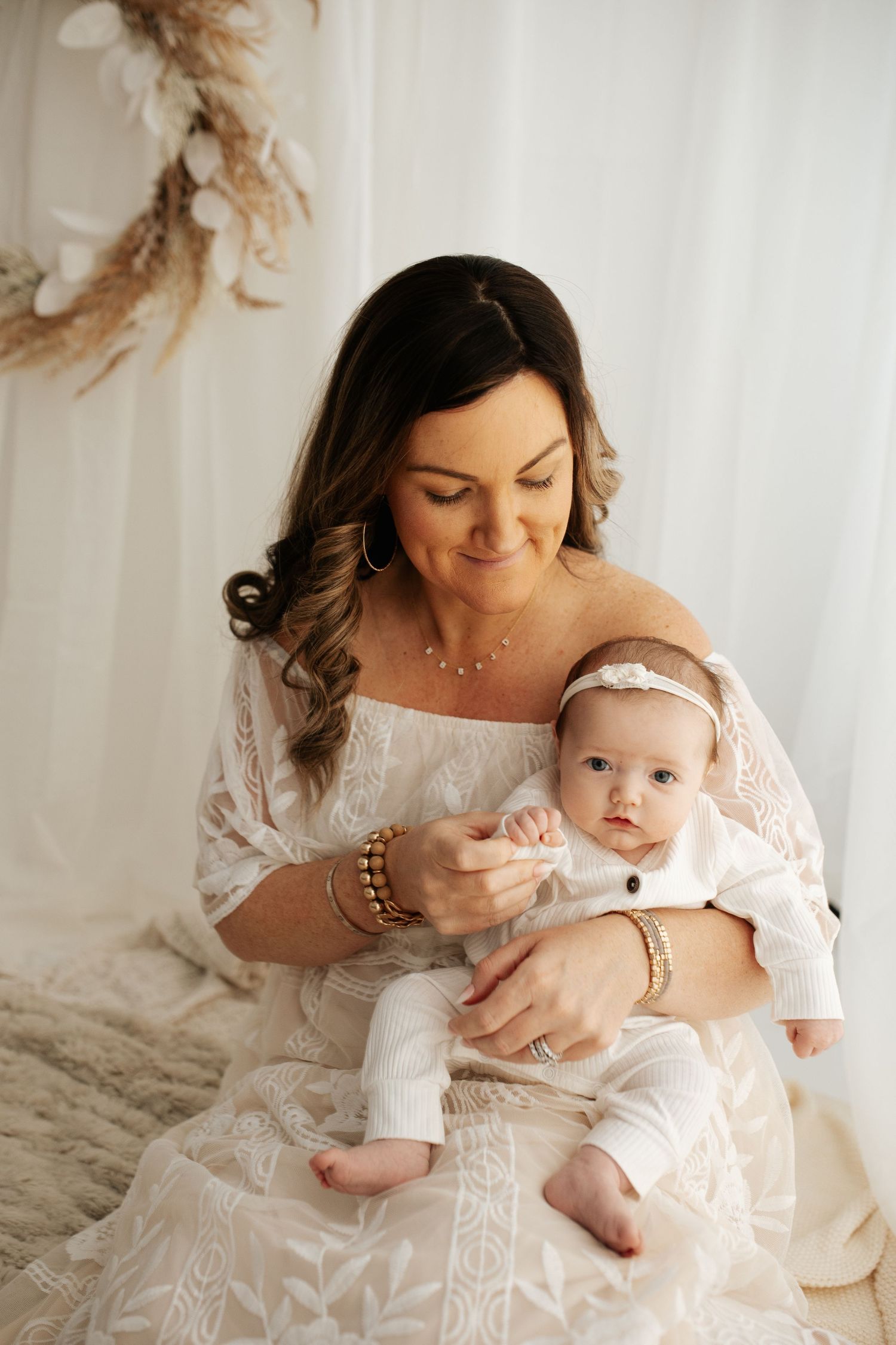 Dark-haired mother in white lace dress holding infant with headband in bright studio setting.