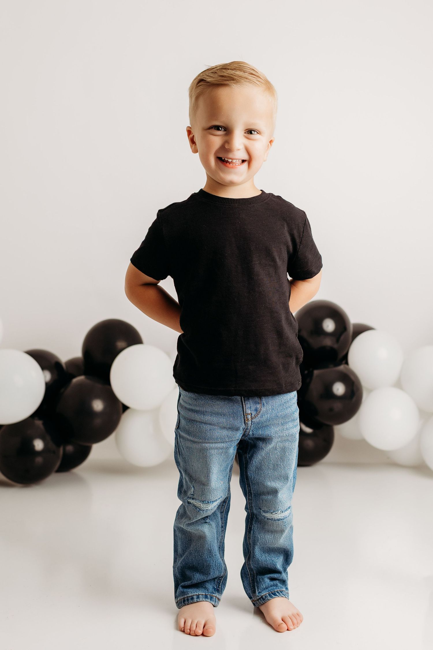 A cheerful young blonde boy in a black t-shirt and jeans poses with black and white balloons.