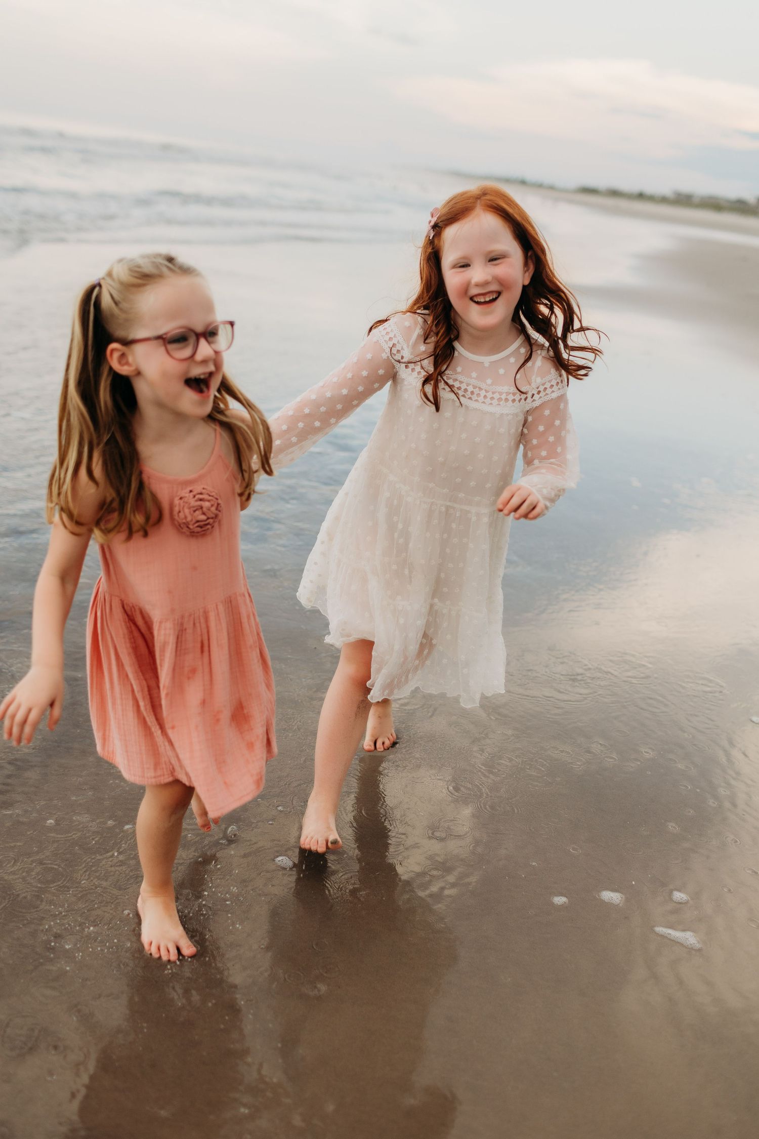 Two laughing girls in pink and white dresses running barefoot along ocean shoreline.