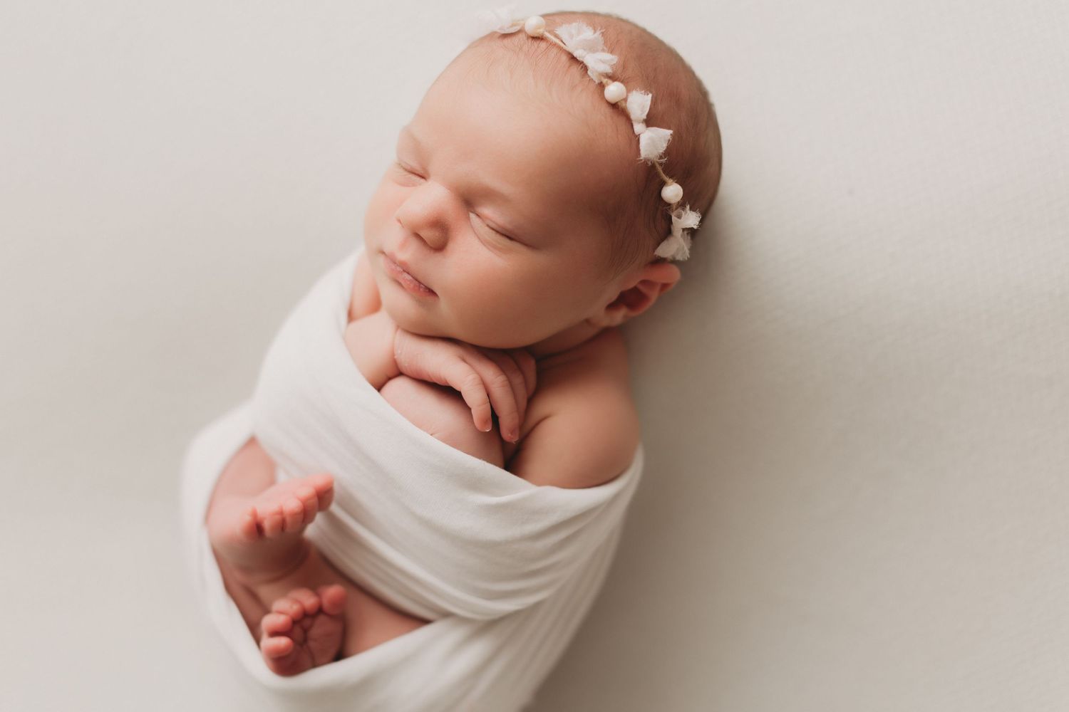 Peaceful sleeping newborn wrapped in soft white fabric wearing delicate white floral headband.