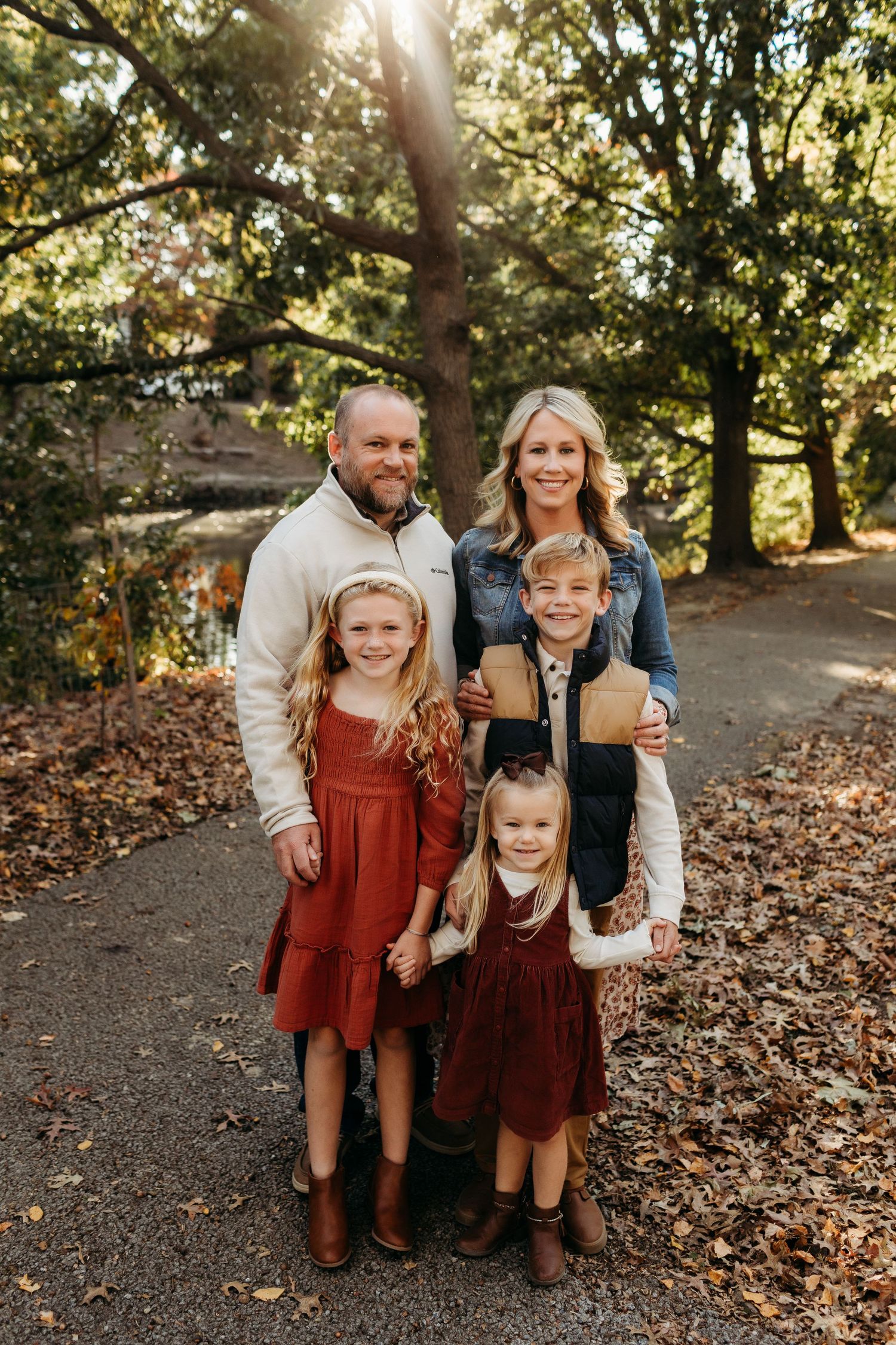Family of five posing on autumn leaf-covered path surrounded by trees in fall portrait.