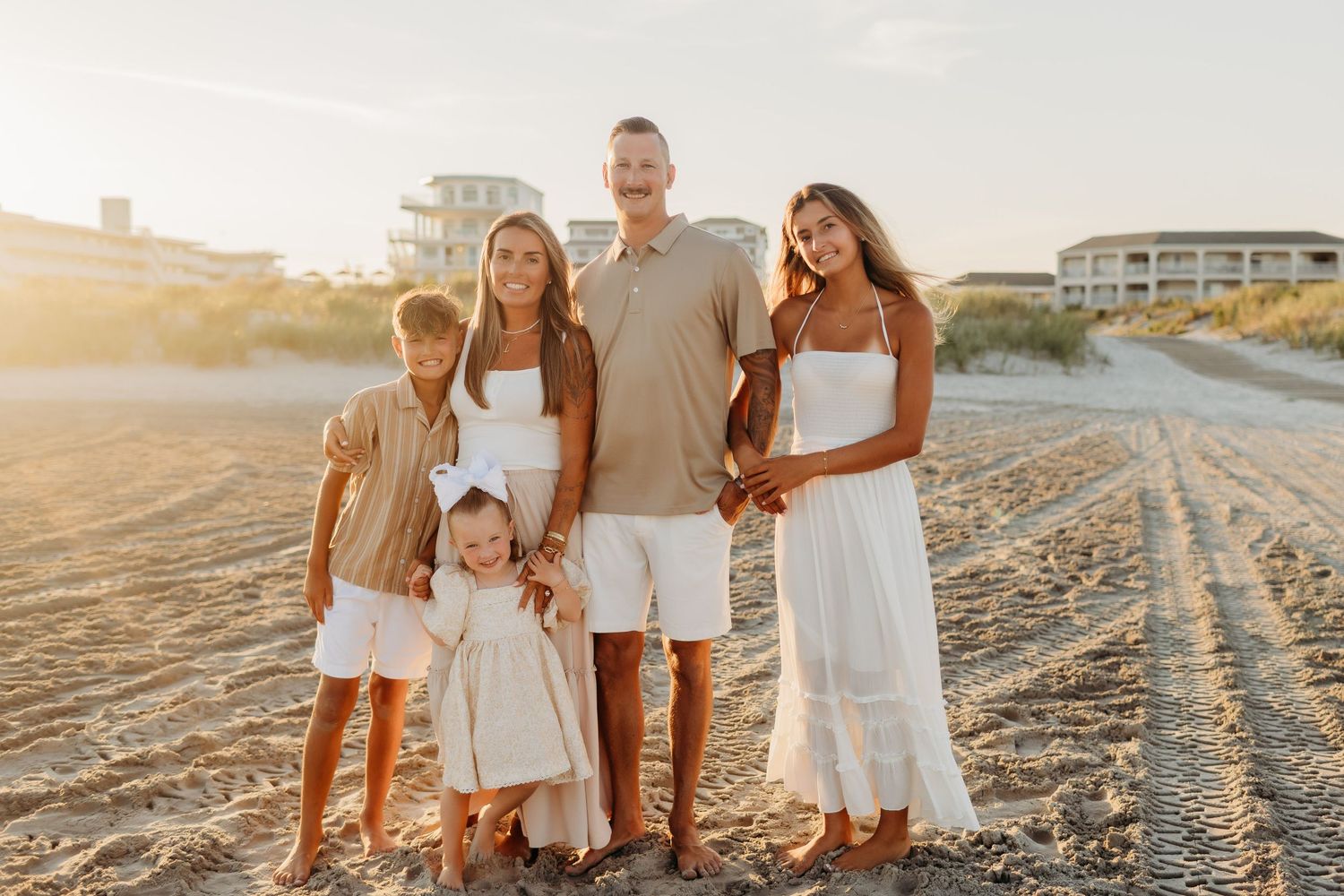 Family of five posing on a sandy beach at golden hour, wearing coordinated neutral and white outfits.