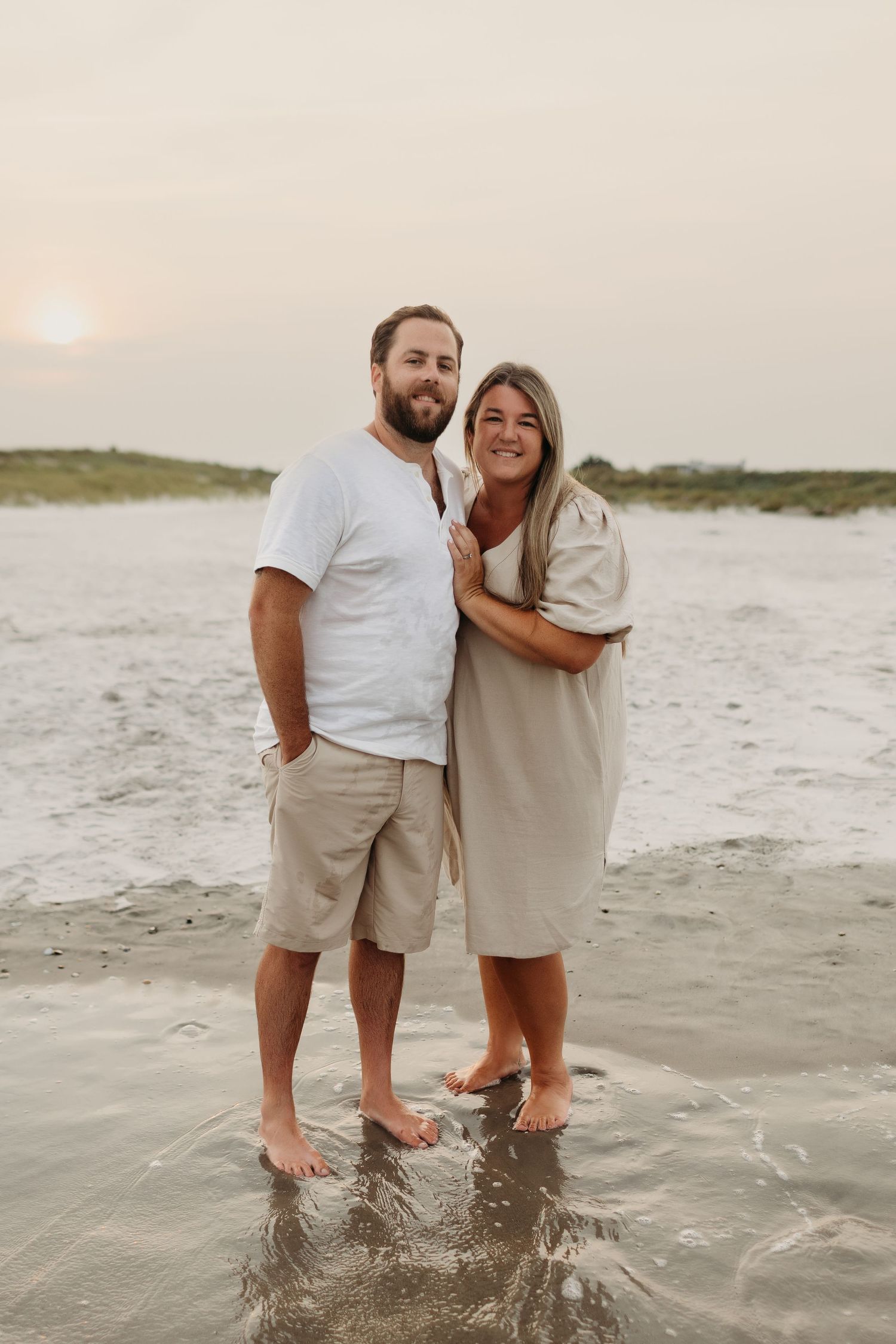 A couple stands barefoot in ocean waves at sunset, the woman in a beige dress embracing the bearded man in a white tee.