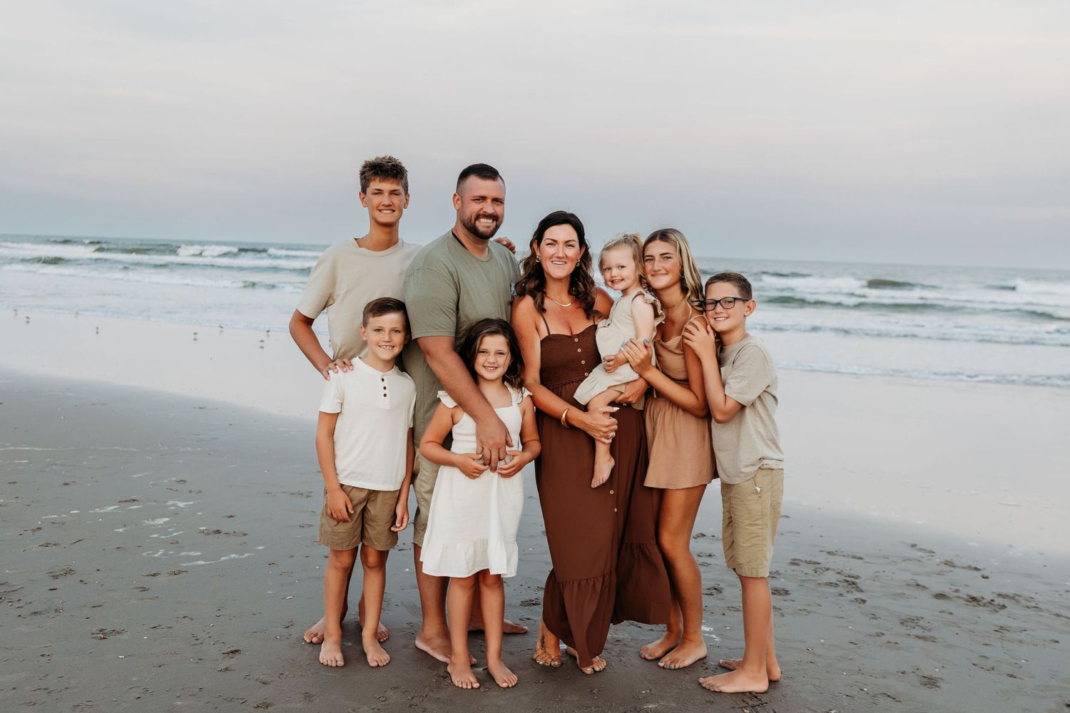 Large family of eight posing together on sandy beach with ocean waves in background.