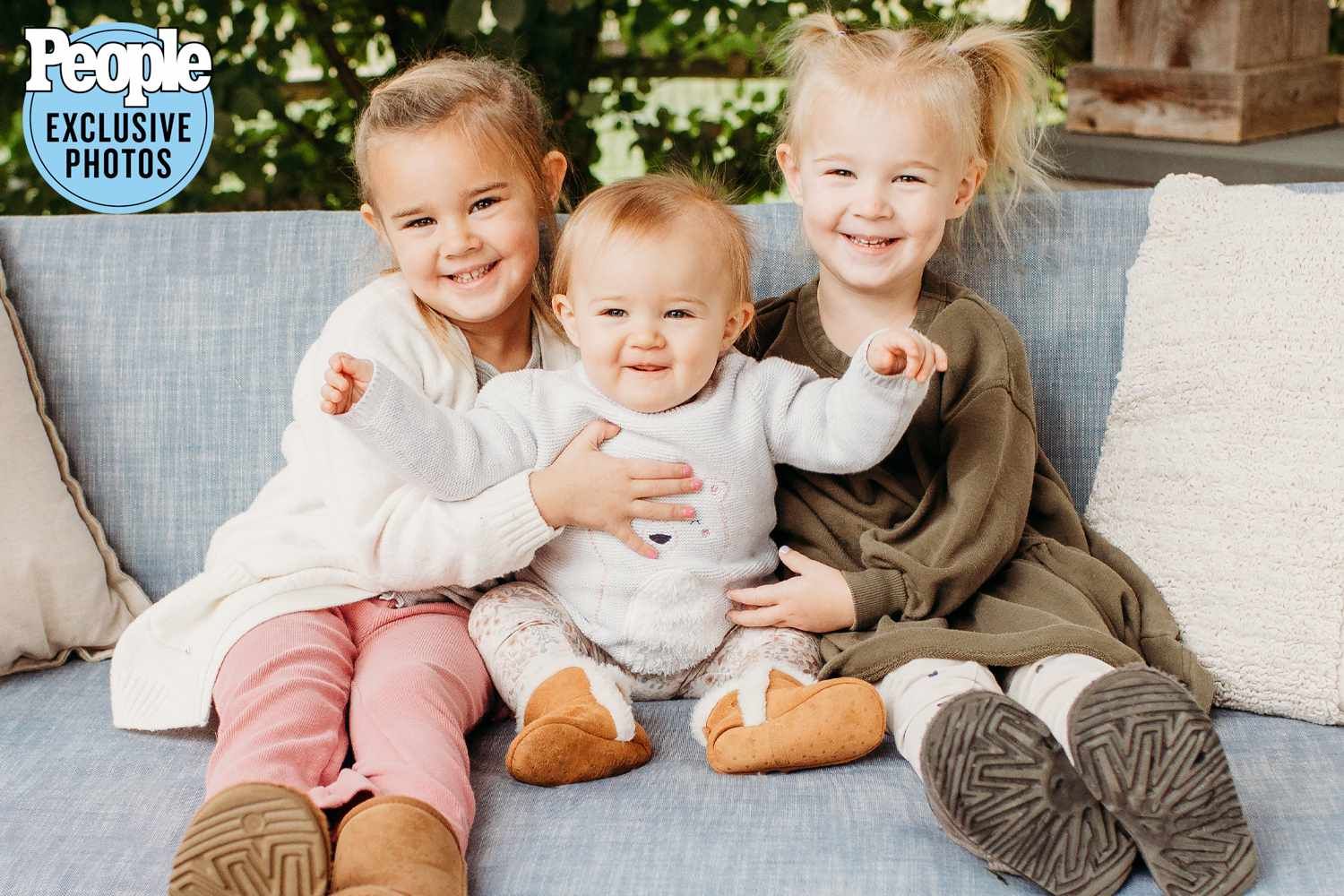 Three young siblings sitting together on couch, two girls hugging baby in white outfit.