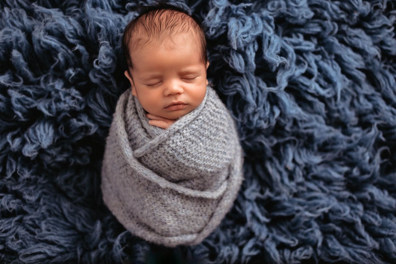 A sleeping newborn wrapped in a cozy gray swaddle blanket on a navy blue textured background.