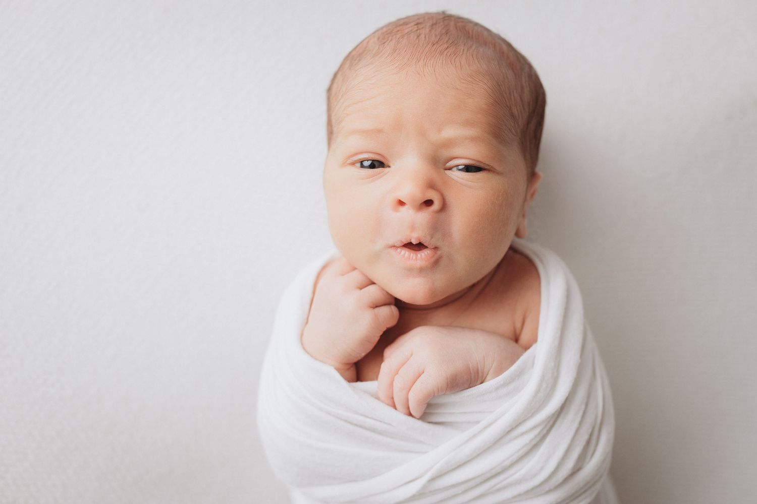 Newborn baby swaddled in white wrap with hand near chin, making an adorable expression against white background.