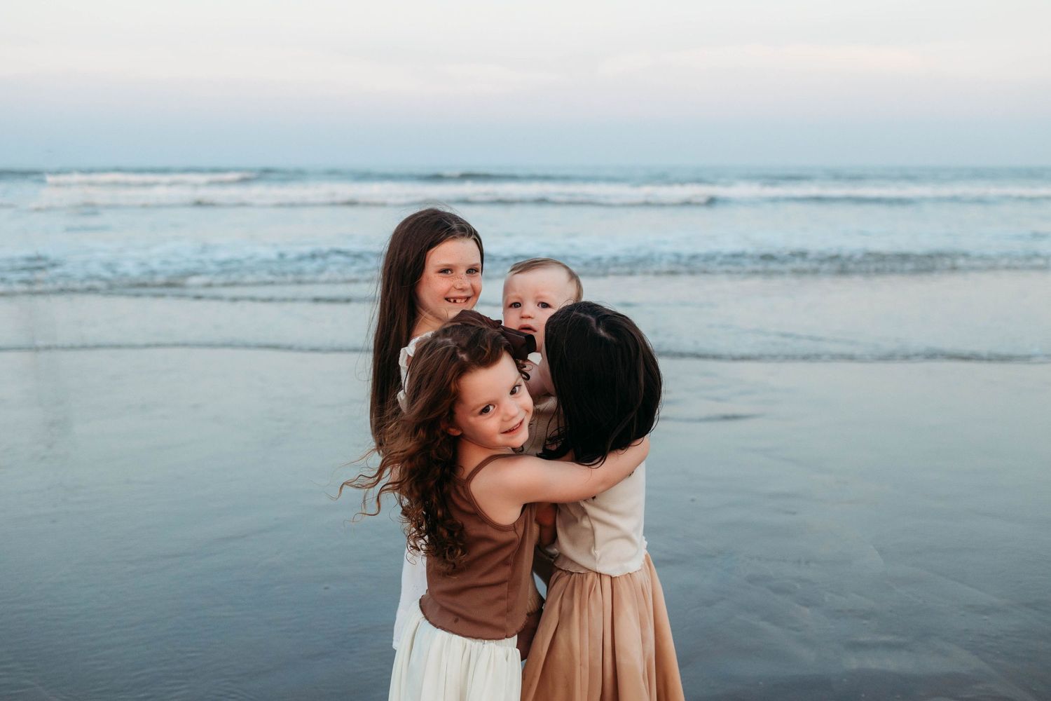 Three people embrace in a warm hug while standing on a misty beach at dusk with ocean waves in the background.