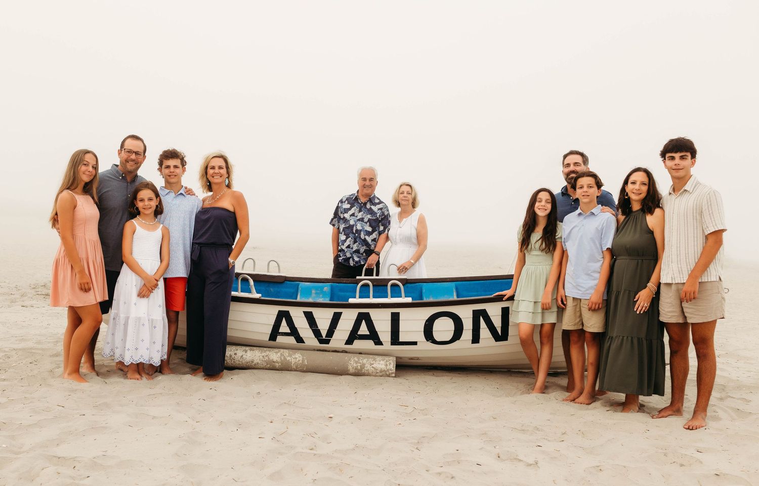 A group of people in casual summer attire pose together on a beach next to a wooden Avalon sign.