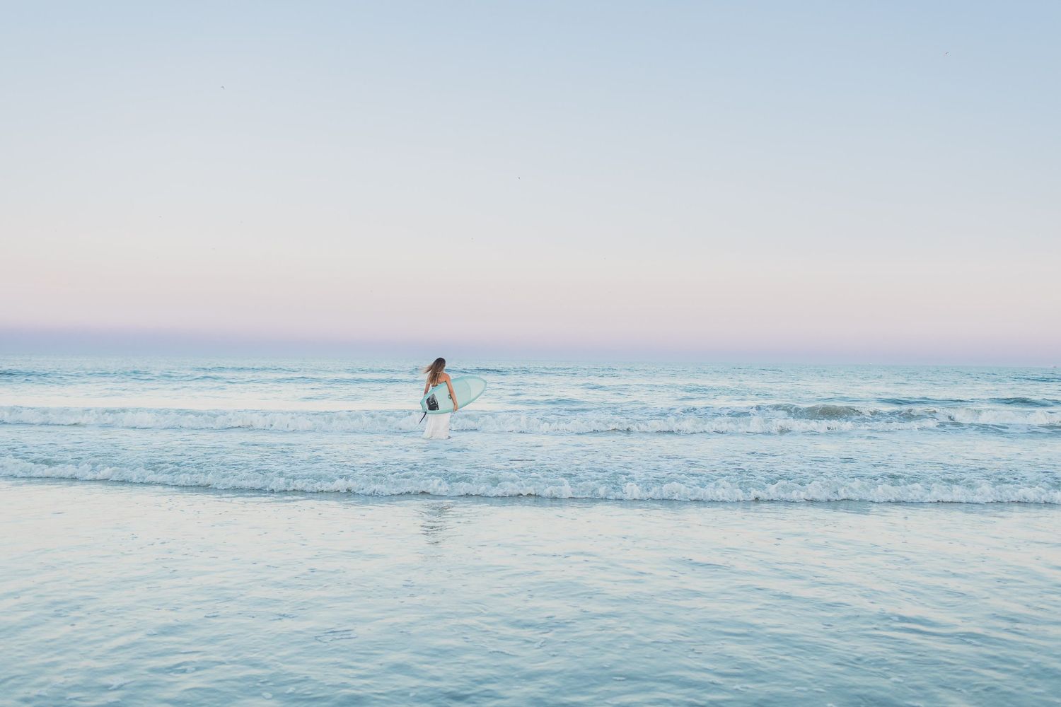 A lone surfer walks into calm blue ocean waves at sunset on a serene beach.