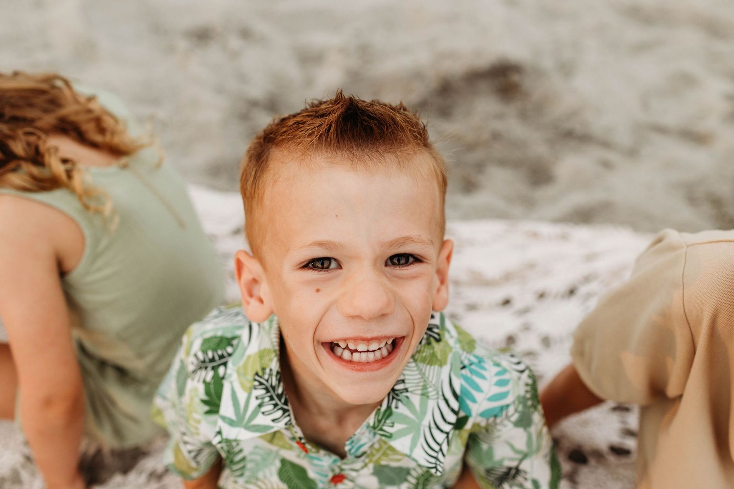 Happy child wearing a tropical print shirt at the beach smiles brightly while sitting on the sand.