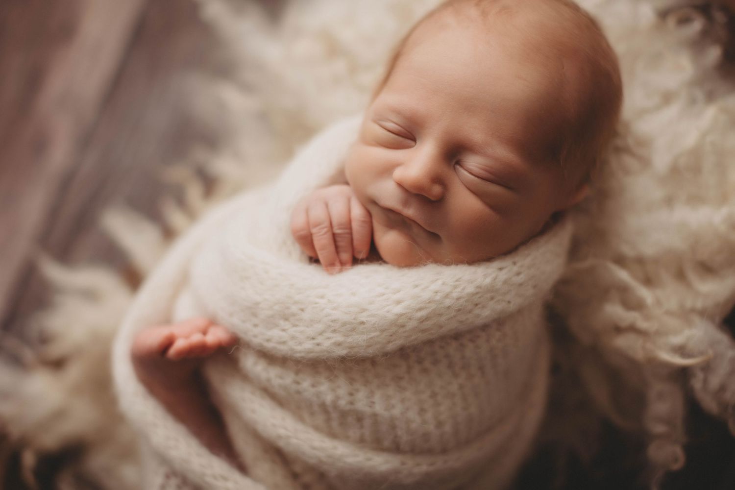 A peaceful sleeping newborn swaddled in a soft white knit wrap rests on cream-colored bedding.