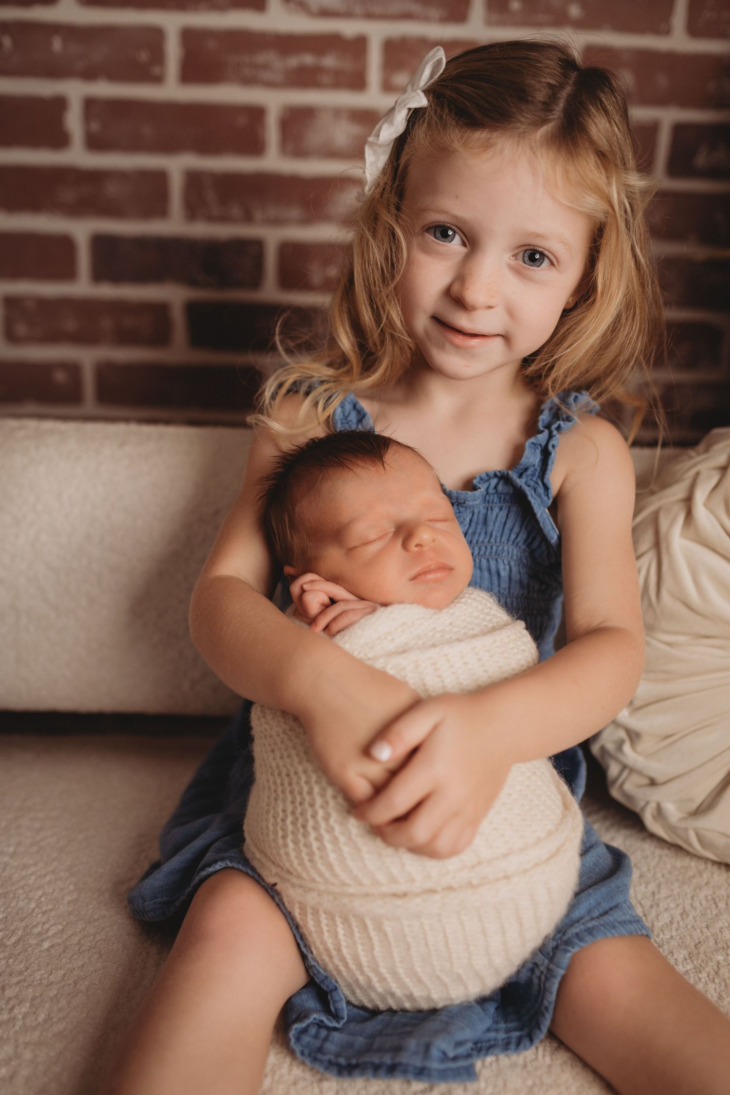 A young girl in a blue dress holds a sleeping swaddled newborn baby while sitting on a white couch near a brick wall.