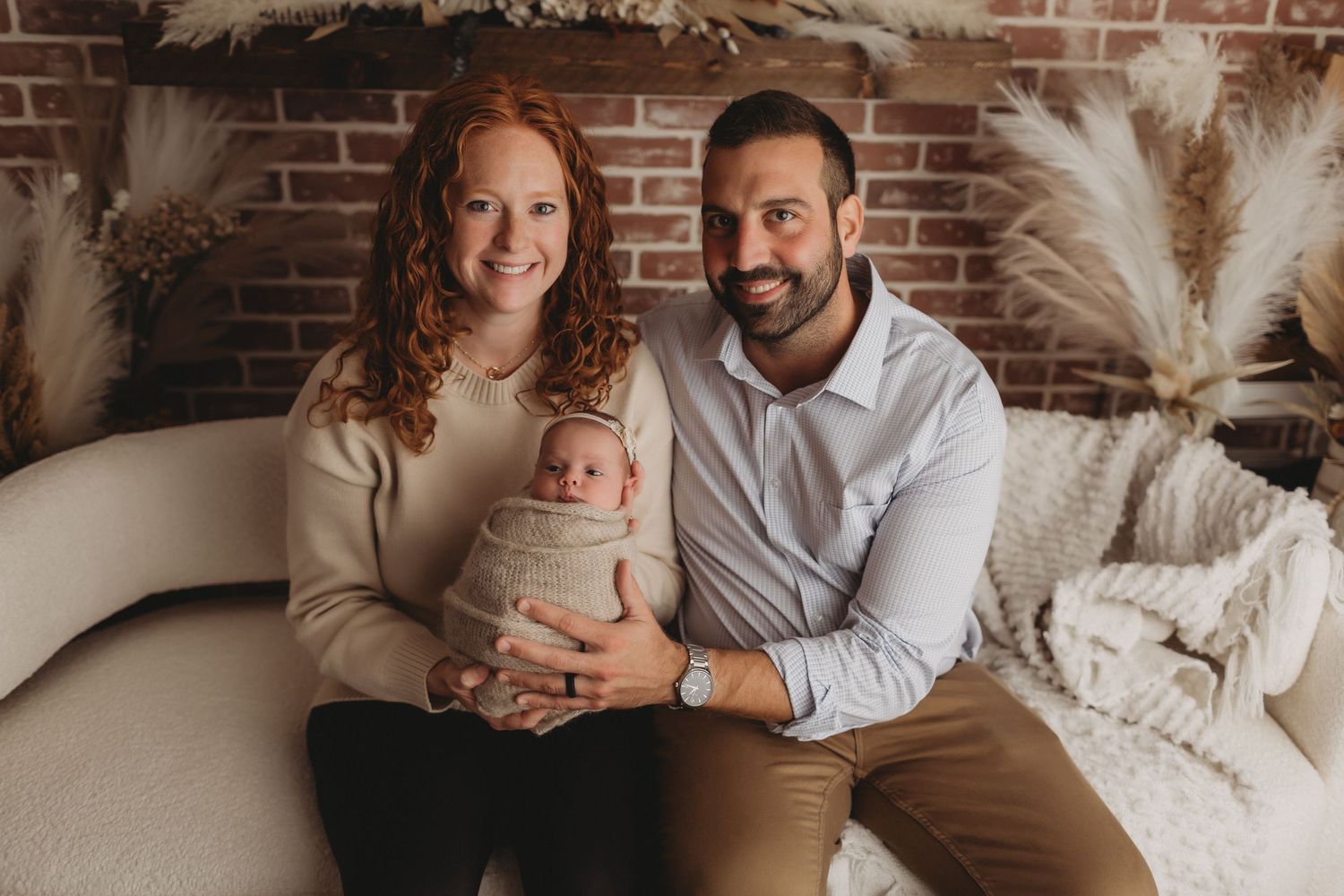 Happy family of three with red-haired mother, dark-haired father, and swaddled newborn posing in studio.