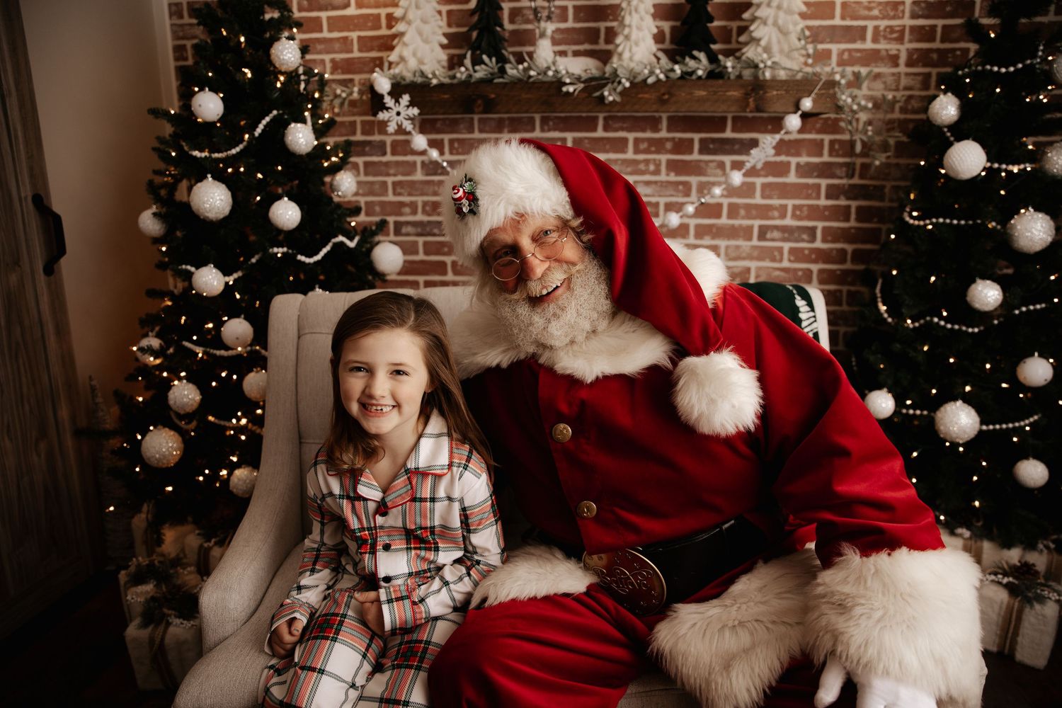 Young girl in plaid pajamas smiling with Santa Claus beside decorated Christmas trees and brick fireplace.