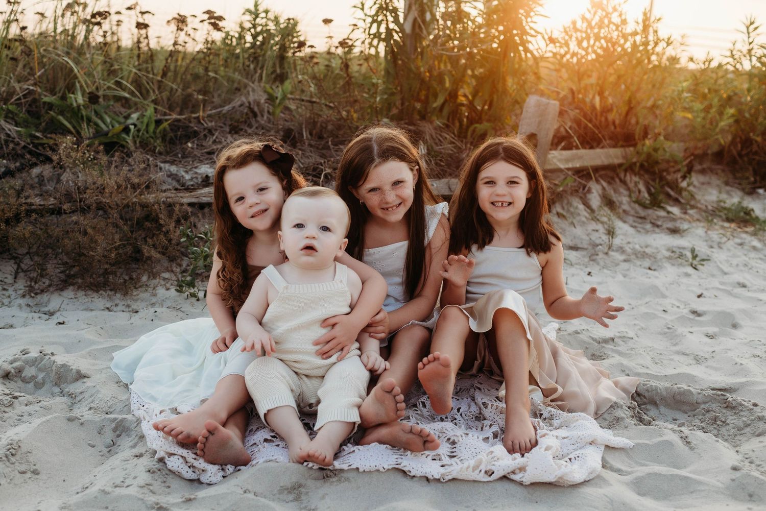 Children in white dresses sit together on a blanket at sunset on a sandy beach.