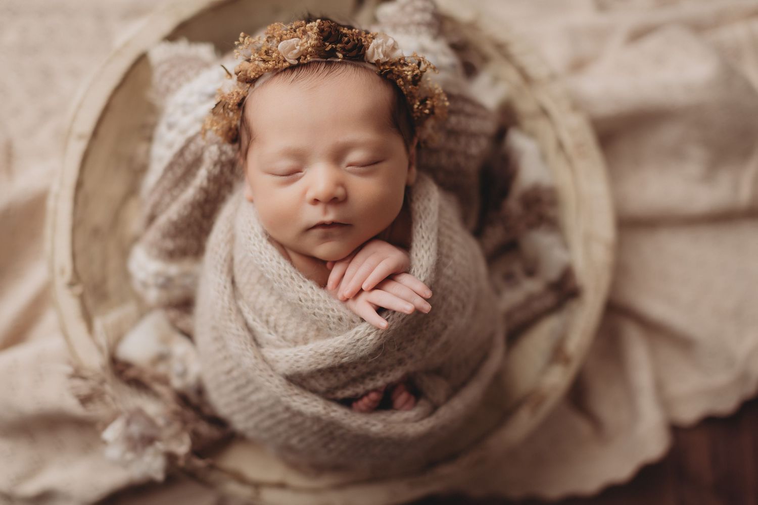 Newborn baby swaddled in beige wrap, sleeping peacefully in a wooden bowl with a floral headband in Ridley Park, PA
