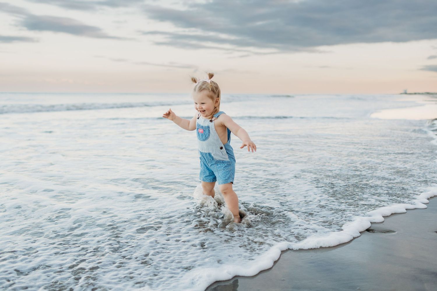 Young child in blue romper joyfully splashing in ocean waves on a sandy beach at sunset.
