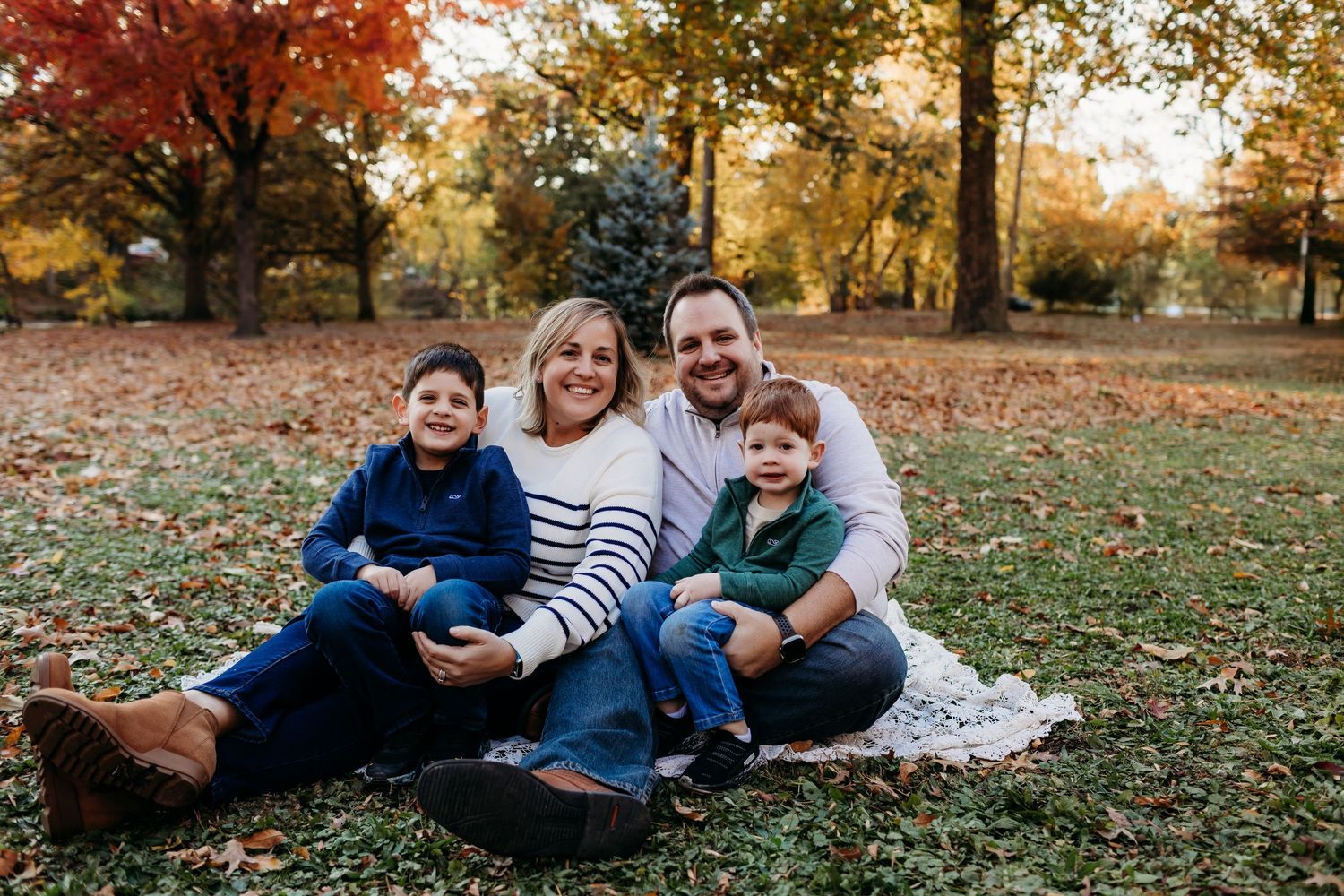 A happy family of four sits on grass surrounded by colorful fall foliage in a park.