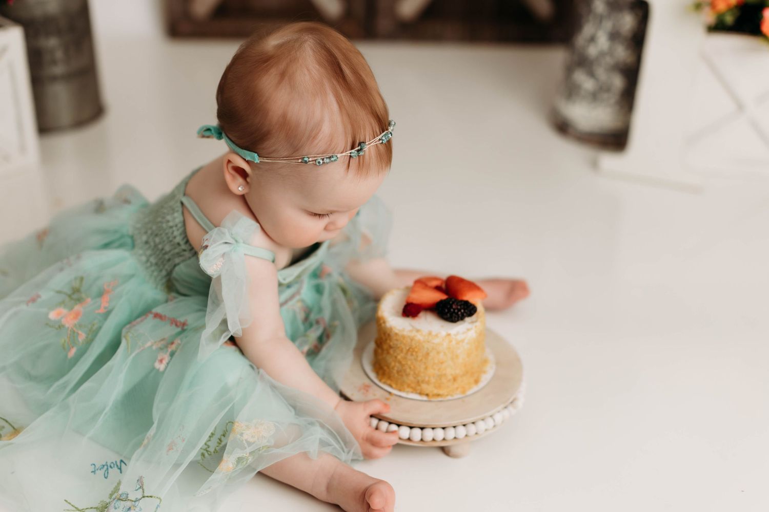 A small child in a mint green tulle dress reaches for a decorated cream cake topped with berries on a white pedestal stand.