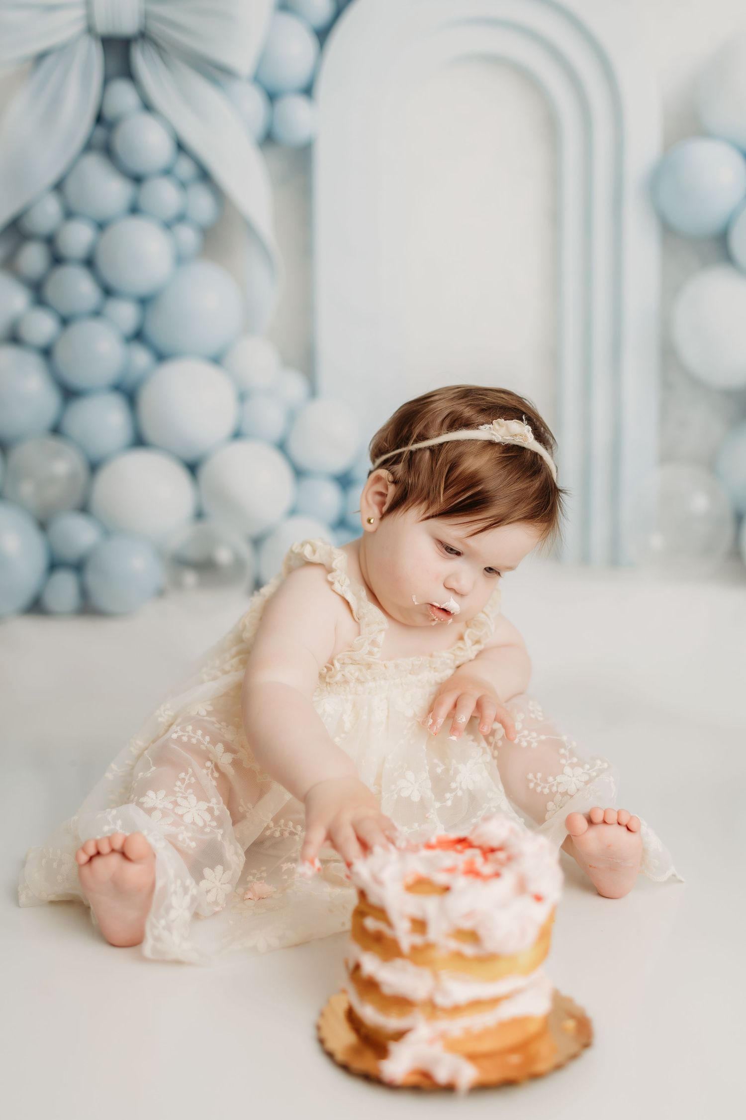 Baby girl in a lace dress reaches for her first birthday smash cake with blue balloon decor.