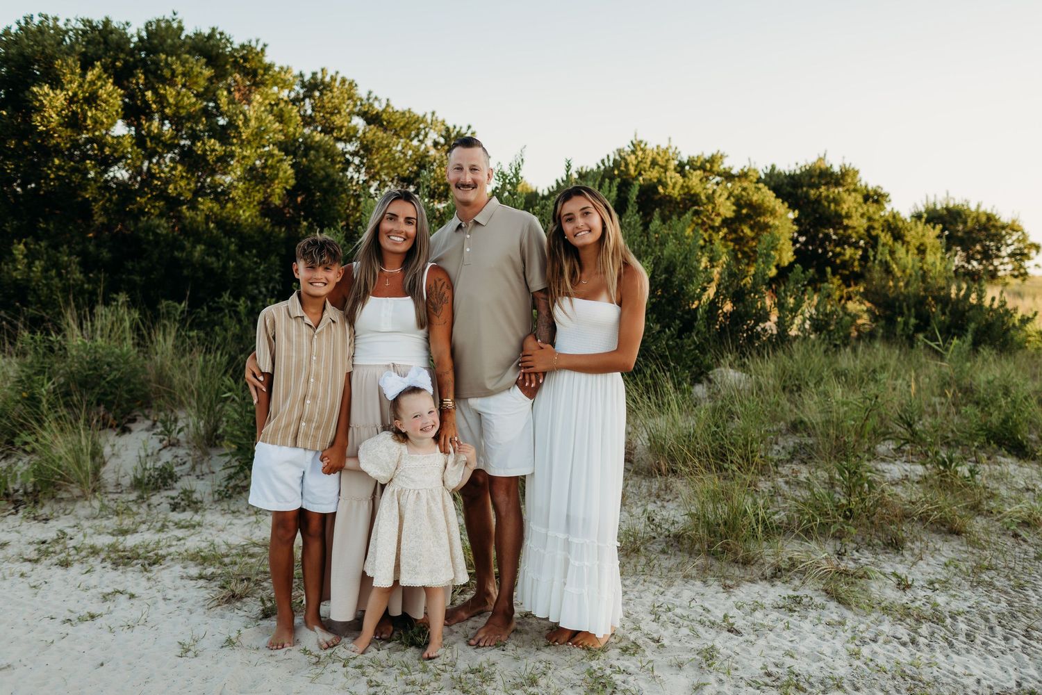 Family of six posing together on a sandy beach with lush green trees in the background during golden hour.