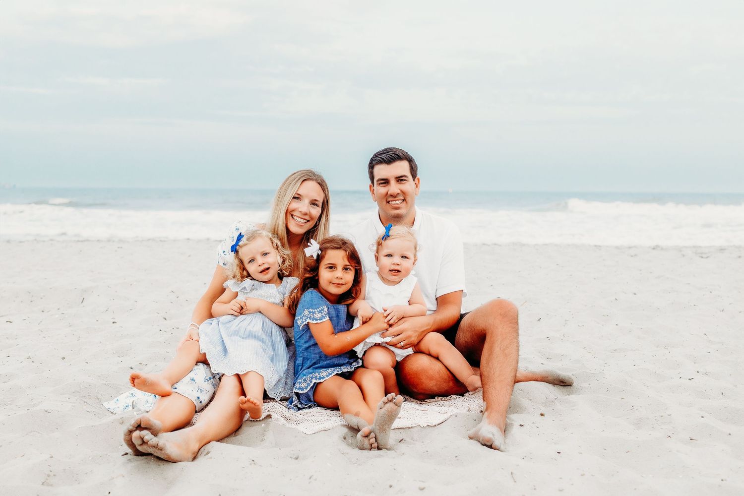 A family sitting together on a sandy beach with ocean waves and cloudy skies in the background.