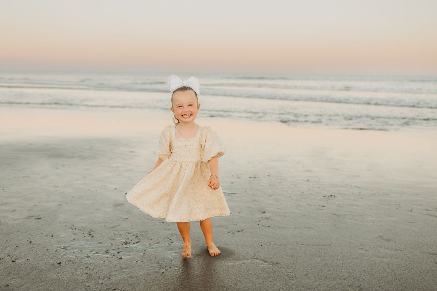 A joyful toddler girl in a cream dress twirls barefoot on a sandy beach at sunset.