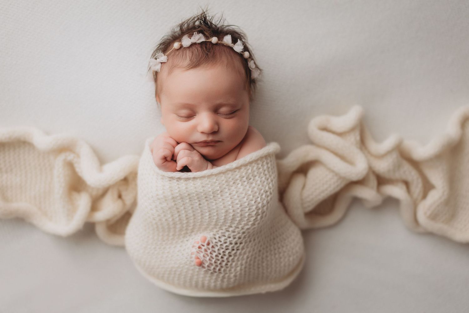 Sleeping newborn baby girl wrapped in cream knit swaddle with floral headband on white background.