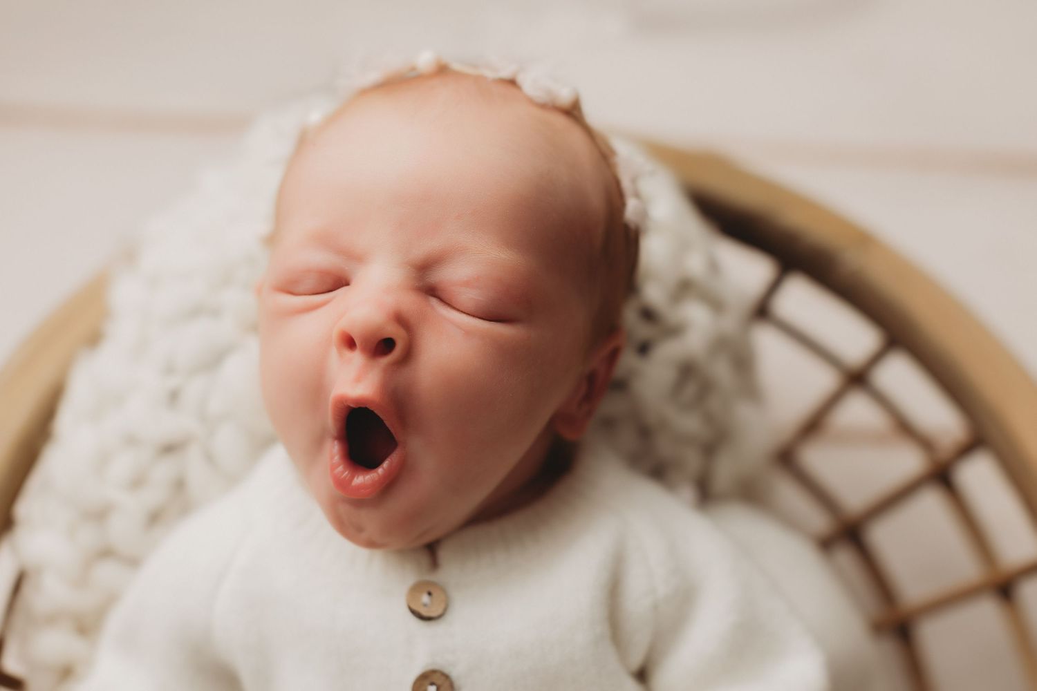 Newborn baby yawning in a wicker basket, wearing a white outfit and floral headband on a soft blanket  in Delaware County studio.
