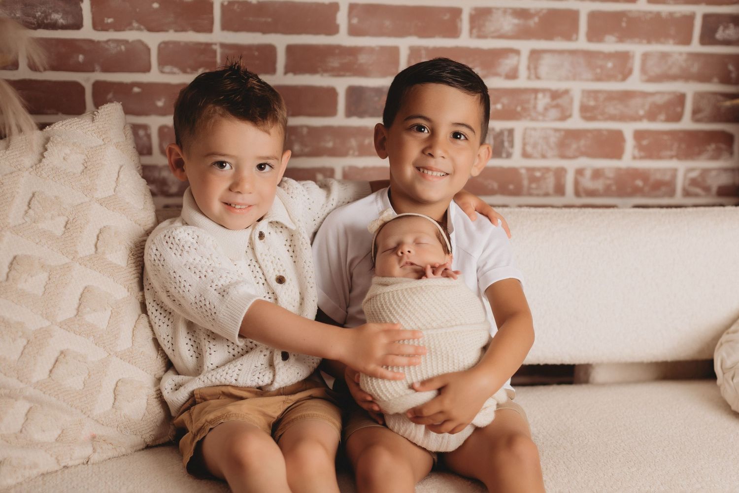Two young brothers smiling on a couch, gently holding their swaddled newborn sibling in a warm brick-wall studio setting.