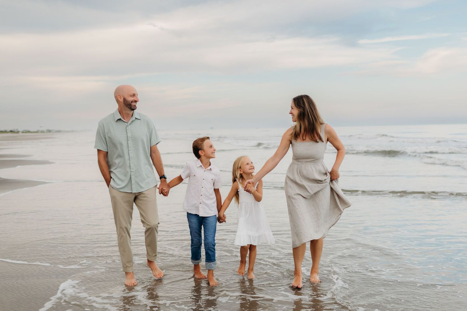 Family of four walking hand-in-hand along a beach shoreline, with waves gently washing over their feet.
