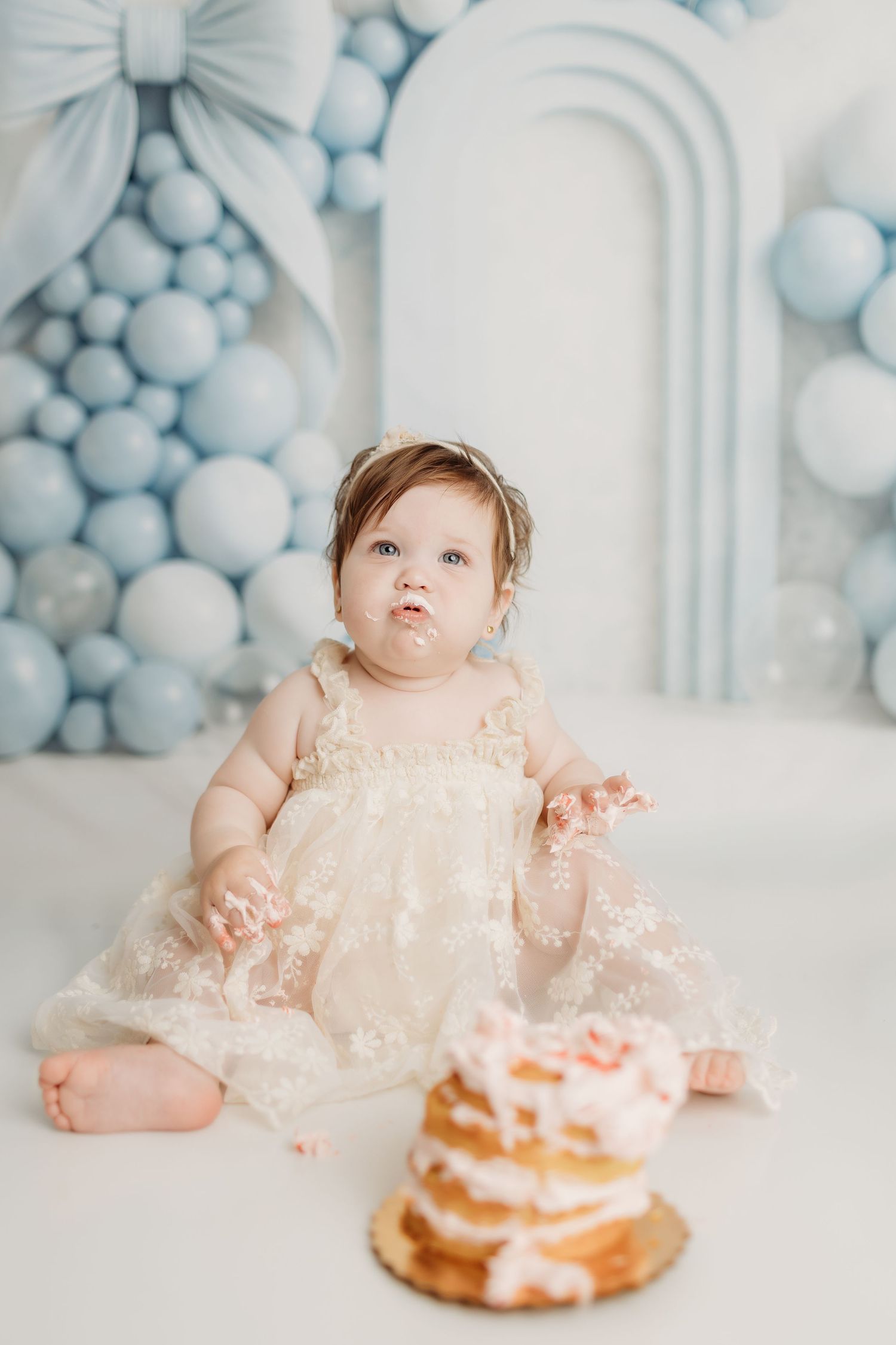 Baby girl in a lace dress sits with cake on her face near a smash cake and blue balloons.