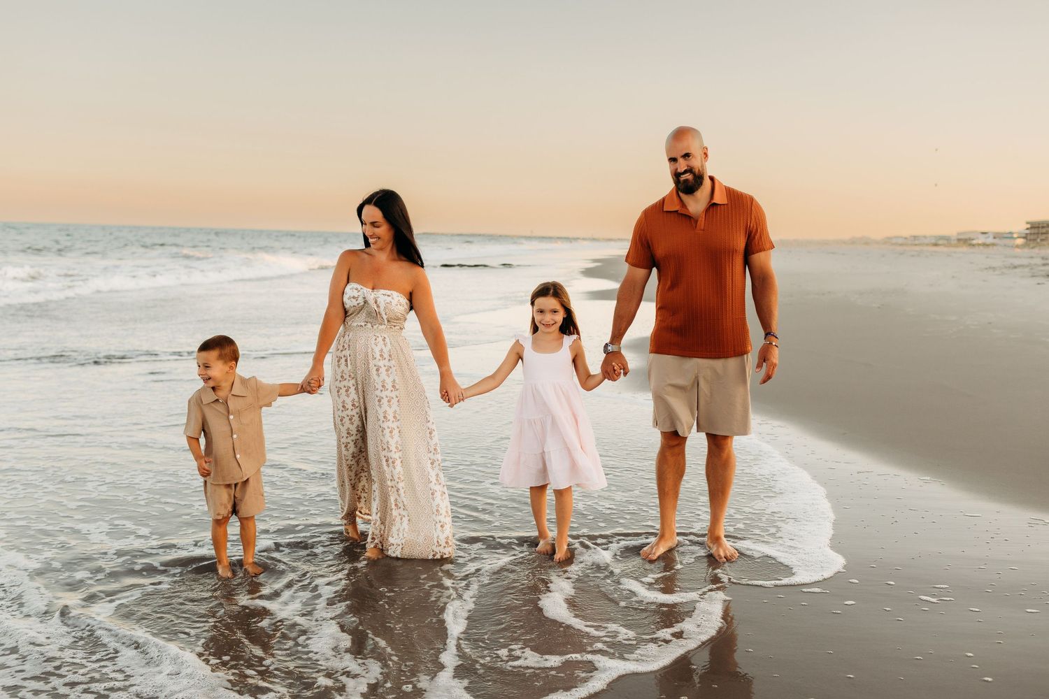 Happy family of four walking along ocean shoreline at sunset in coordinated outfits.
