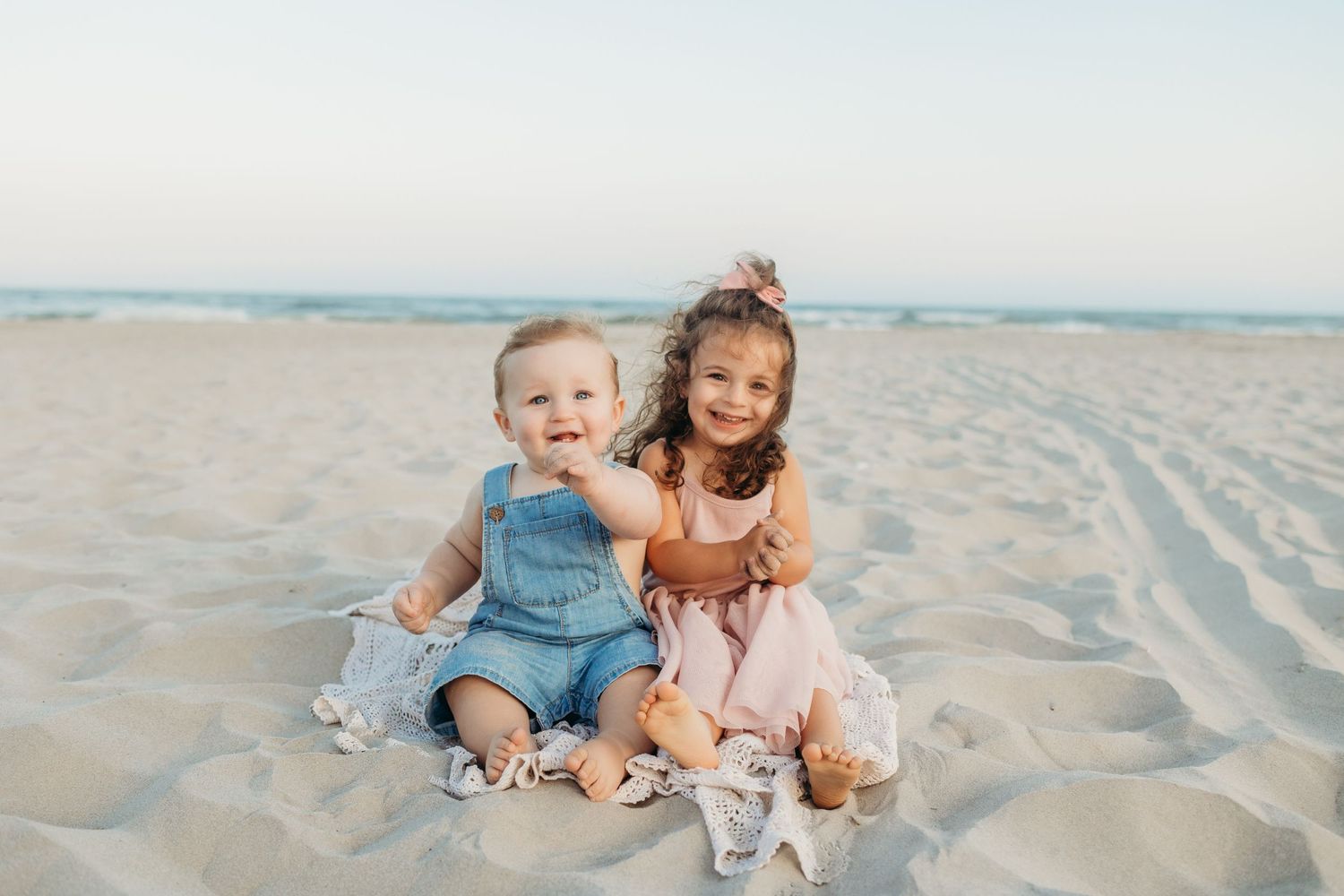 Two young children sitting together on a sandy beach during a peaceful sunset.