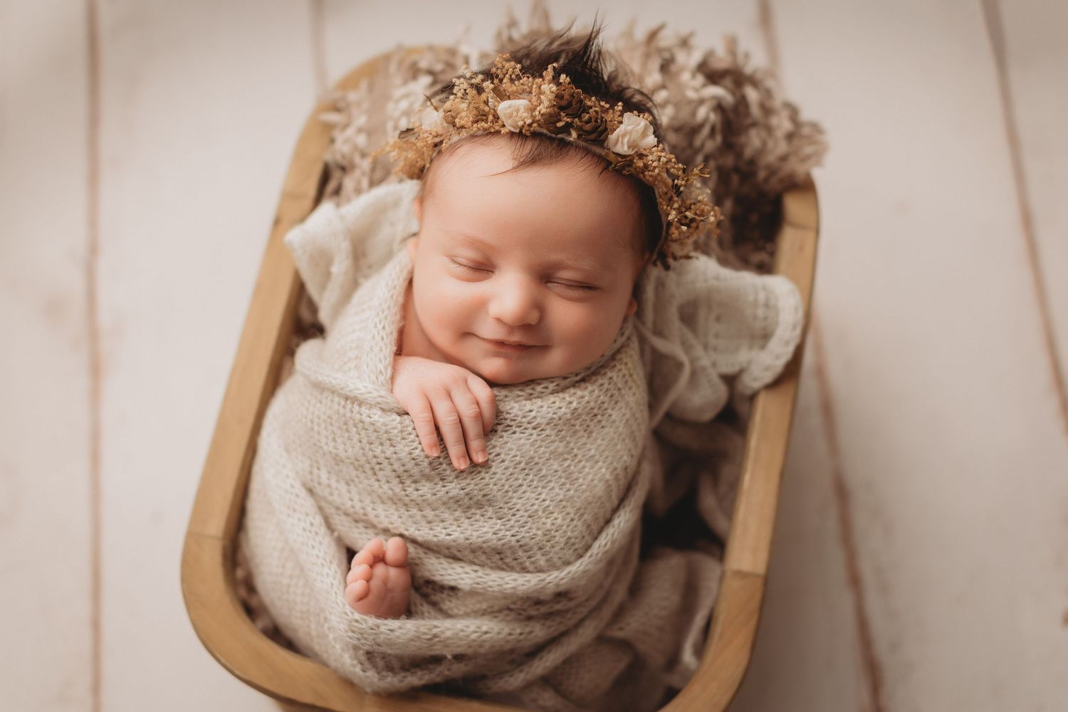 Smiling newborn baby swaddled in beige wrap wearing floral crown, sleeping peacefully in wooden bowl prop.