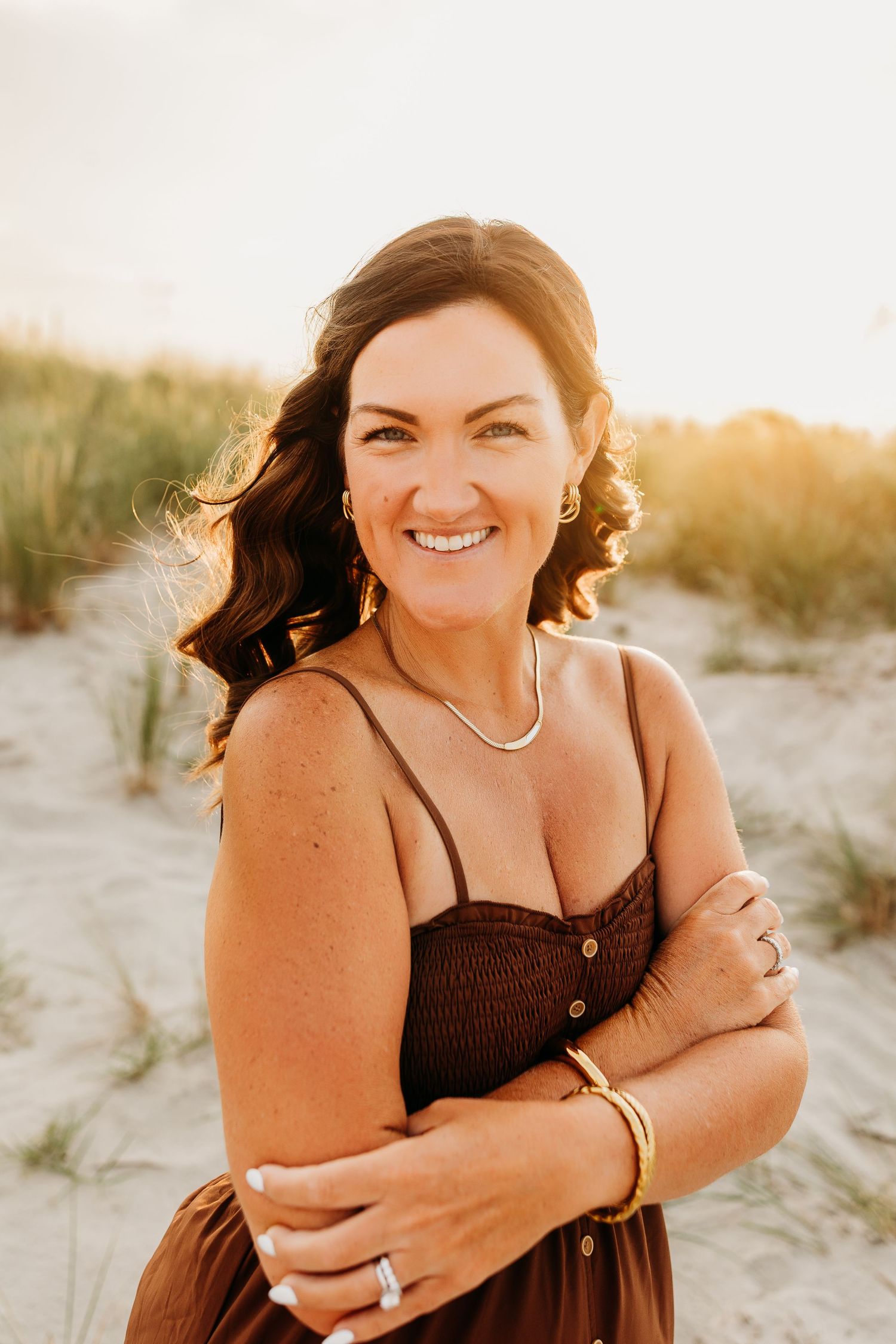 Smiling brunette woman in brown dress posing on sandy beach dunes at golden sunset.